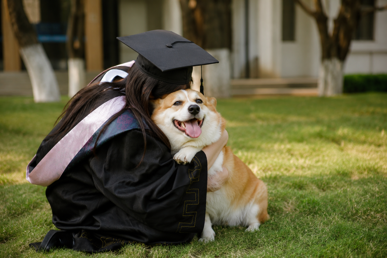 A graduate in a cap hugging her happy Corgi on a grassy lawn after the ceremony
